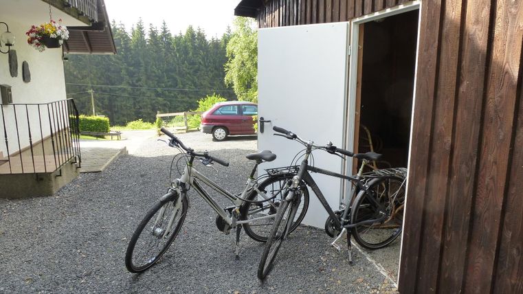 Twee fietsen staan naast een open deur van een houten gebouw. Op de achtergrond is een auto en een groene landschaps te zien.