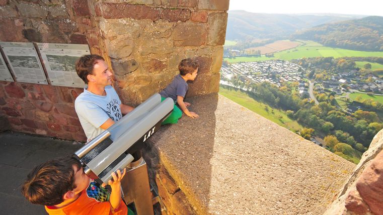 A father and two children are looking through a telescope from an elevated spot. In the background, the beautiful landscape with a village and hills can be seen.