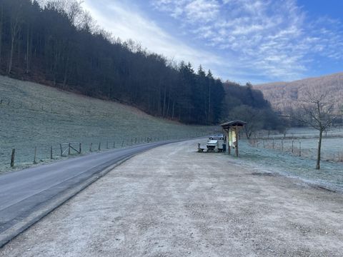 A quiet country road with frost on the ground and trees in the background. A small waiting area is located at the edge of the road.