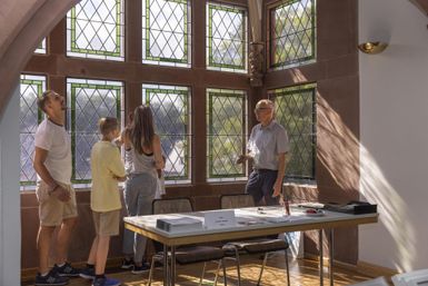 Eine Gruppe von vier Personen steht vor einem großen, mit bunten Fenstern ausgestatteten Raum. Ein älterer Mann erklärt etwas, während die anderen aufmerksam zuhören.