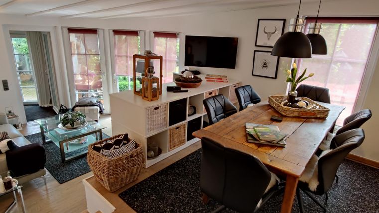Een gezellige woonkamer met een eettafel en een moderne bank. Grote ramen laten veel licht binnen, en decoratieve elementen creëren een uitnodigende sfeer.
