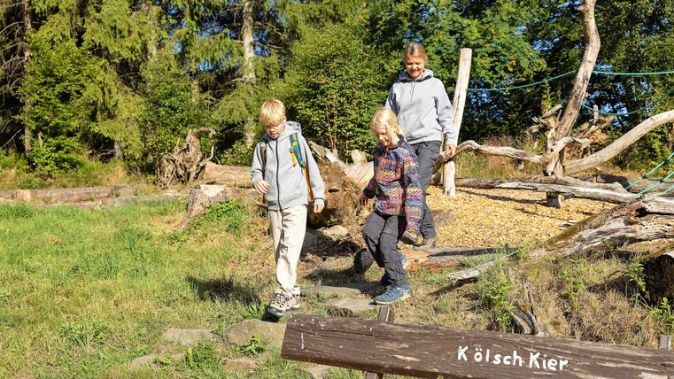 Three children are hiking on a forest path. In the foreground, there is a wooden sign with the inscription "Köläch Kier".