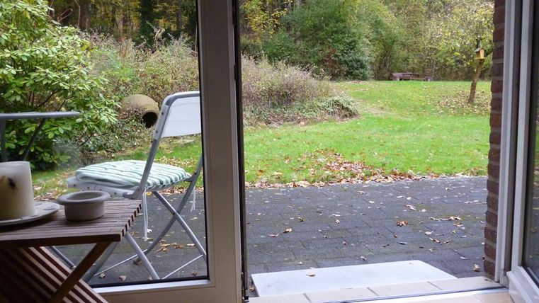 Een blik uit een raam op een tuin met gras en struiken. Op de voorgrond staat een stoel en een tafel.