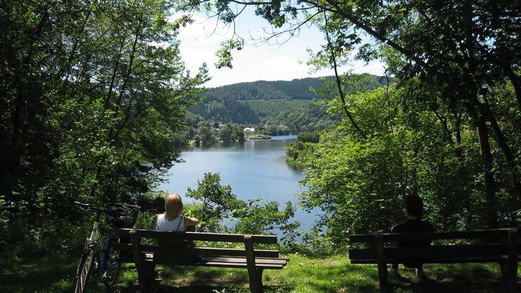 A tranquil lake surrounded by trees and hills. Two people are sitting on benches and enjoying the view.