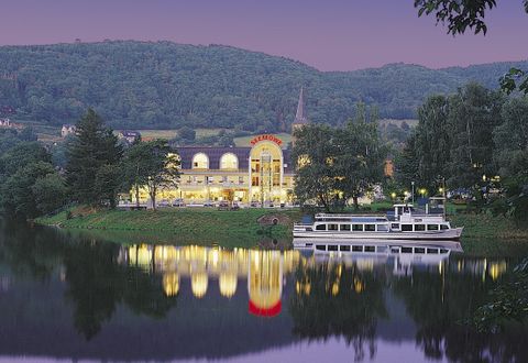 A picturesque view of a building by the river with gentle hills in the background. The light from the building is reflected on the water.