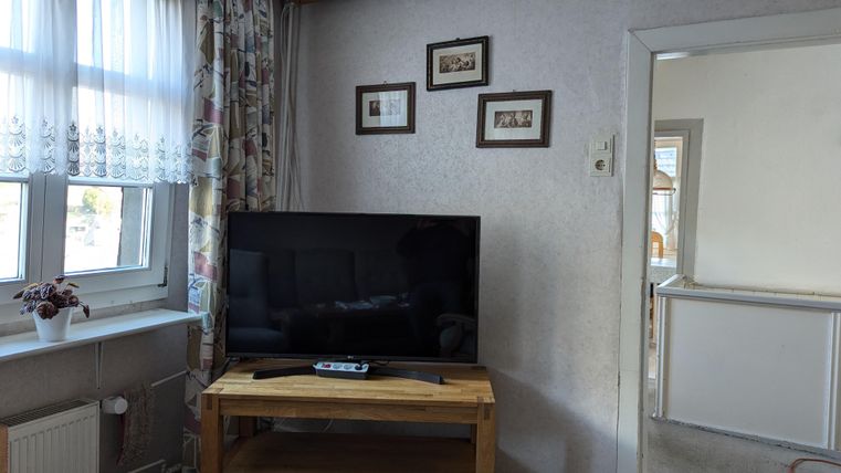 A living room with a television on a wooden stand. On the wall hang some pictures and a window with curtains is also visible.