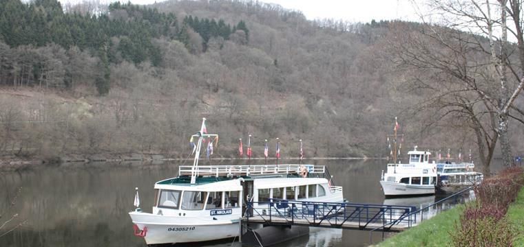 Twee excursieboten liggen aan een steiger bij de rivier. Op de achtergrond zijn beboste heuvels te zien.