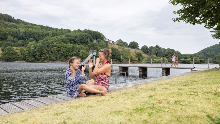 Twee meisjes spelen en lachen aan de oever van een waterlichaam. Op de achtergrond is een houten brug en groene heuvels te zien.