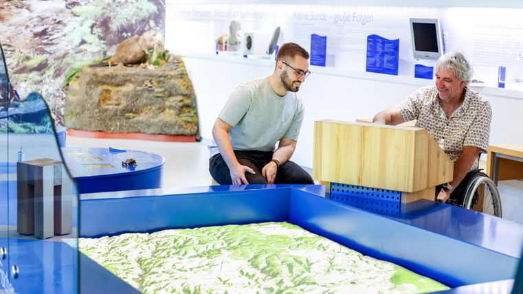 Zwei Männer arbeiten an einem interaktiven Modell in einem Ausstellungsraum. Das Modell zeigt eine topografische Landschaft und die Männer scheinen konzentriert und interessiert zu sein.