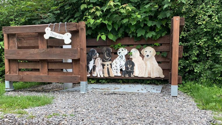A wooden dog play station with various dog silhouettes. In the background, there is a green area with plants visible.