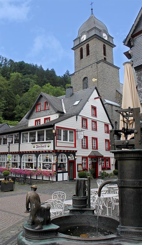 A picturesque village street with traditional half-timbered houses and a church tower in the background. In the foreground, there is a fountain with a childlike statue.