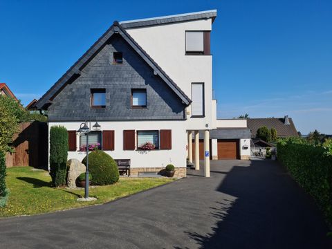 Ein modernes Haus mit einem schrägen Dach und großen Fenstern. Der Eingangsbereich ist von grünen Pflanzen umgeben und die Auffahrt ist gepflastert.
