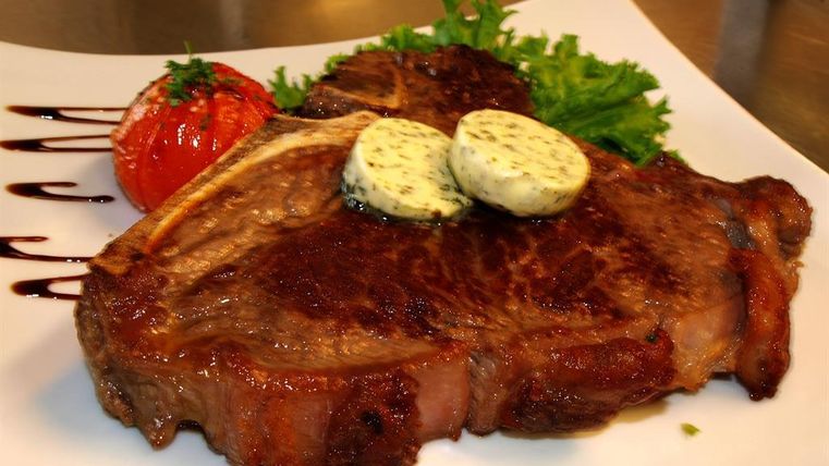 Een sappige steak met kruidenboter, geserveerd met een gebakken tomaat en verse salade. Het bord is elegant opgemaakt en ziet er smakelijk uit.