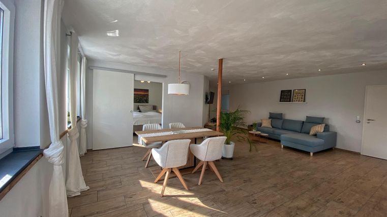 A bright and modern living and dining room with a large table and white chairs. The room has a cozy atmosphere with a blue couch and large windows.