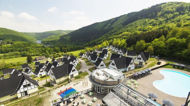 Eine malerische Ferienanlage inmitten von grünen Hügeln. Zu sehen sind gemütliche Häuser und ein Poolbereich mit Liegen.