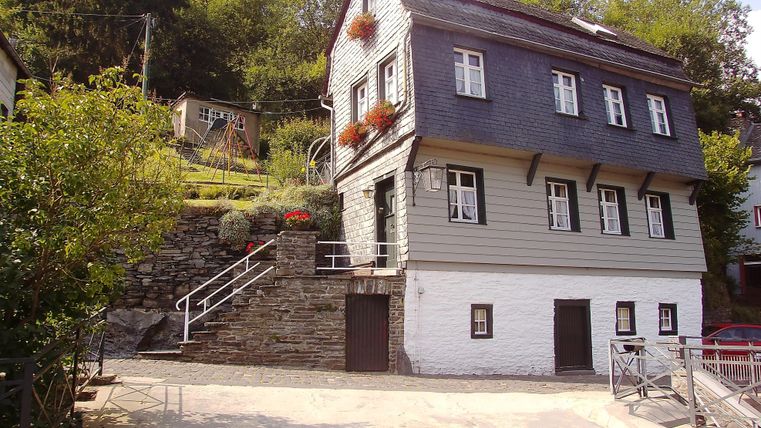 A historic house with a gray facade and white accents. It is situated in a green environment with steps and a small garden.