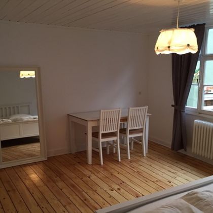 A simple and bright room with a table and four chairs. A large mirror is mounted on the wall and the windows let in plenty of daylight.