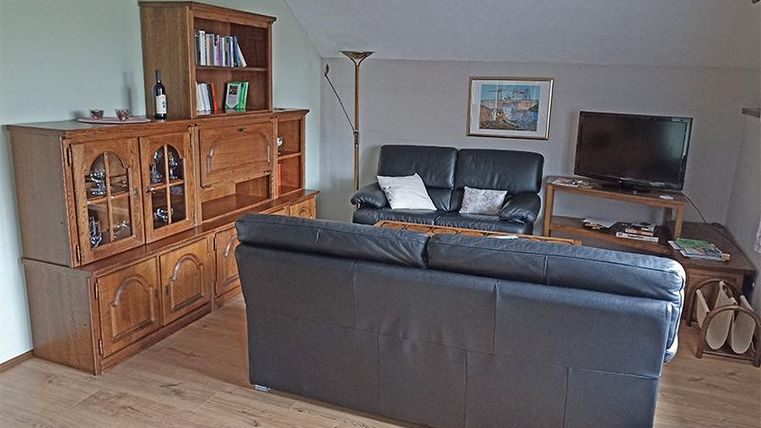 Ein Wohnraum mit einem schwarzen Sofa und einer hellen Holzvitrine. In der Ecke steht ein Fernseher und an der Wand hängt ein Bild.
