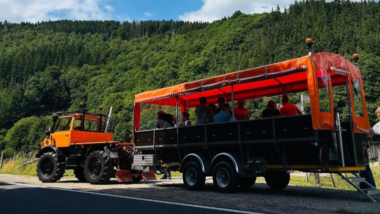 Een tractor trekt een wagen met veel passagiers. Het uitzicht is groen en bergachtig, met een blauwe lucht op de achtergrond.