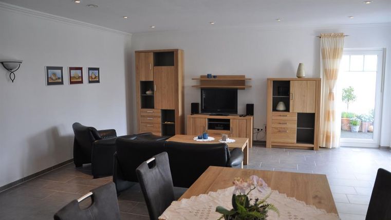 Ein modernes Wohnzimmer mit einer gemütlichen Sitzecke und einem Fernseher. An einem Tisch steht eine Blumendekoration, und es gibt große Fenster mit Licht.
