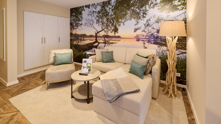 Ein modernes Wohnzimmer mit einem bequemen Sofa und einem stilvollen Lampe. Im Hintergrund ist eine Fototapete mit einer idyllischen Landschaft bei Sonnenuntergang.