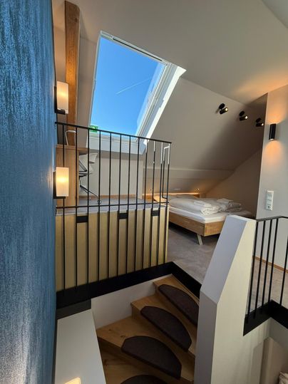 Een moderne interieurruimte met houten trap en helder licht. Boven bevindt zich een gezellige slaapkamer met een groot raam.