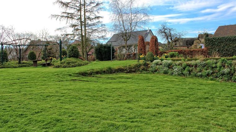 Een onderhouden tuin met groene gras en kleurrijke bloemen. Op de achtergrond zijn enkele huizen en bomen te zien.