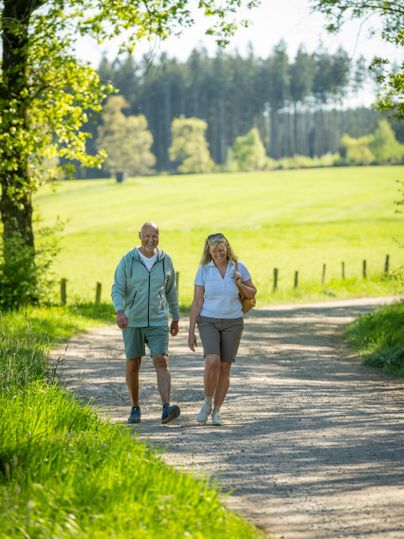Ein Paar spaziert auf einem Naturweg in einer grünen Landschaft. Die Sonne scheint und es ist ein schöner, klarer Tag.
