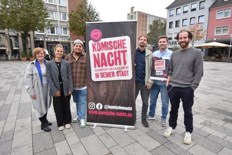 Eine Gruppe von sechs Personen steht vor einem Banner, das für eine Comedy-Veranstaltung mit dem Titel "Komische Nacht" wirbt. Die Szenerie ist urban, mit modernen Gebäuden im Hintergrund.