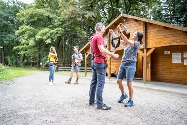 Eine Gruppe von Menschen bereitet sich auf ein Kletterabenteuer vor. Sie stehen an einer Holzhütte im Wald und tauschen Kletterausrüstung aus.