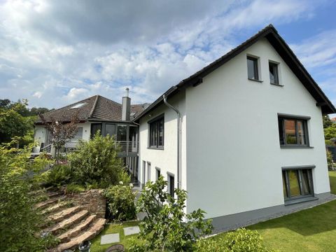 A modern house with white walls and a bright roof. The garden is green and well-kept with a stone staircase.