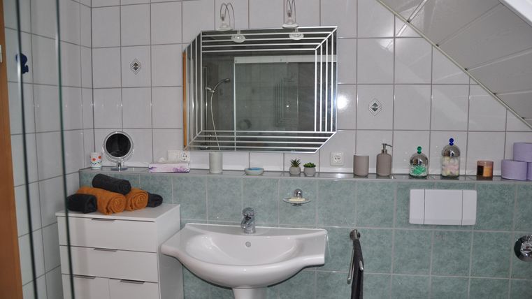 A modern bathroom with green tiles. There is a sink, a mirror, and various toiletries on the shelves.