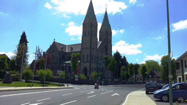 Eine beeindruckende Kirche mit zwei hohen Türmen vor blauem Himmel. Die Umgebung ist grün und einladend, mit einer Straße im Vordergrund.