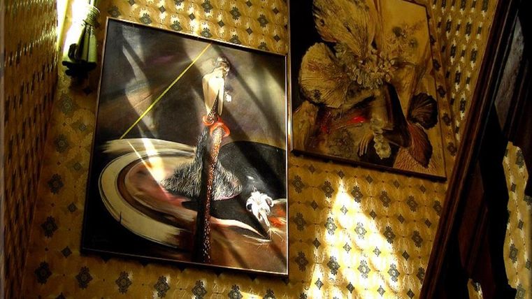 Zwei Kunstwerke an einer Wand mit einem gemusterten Hintergrund. Das Licht fällt sanft auf die Bilder und schafft eine interessante Atmosphäre.