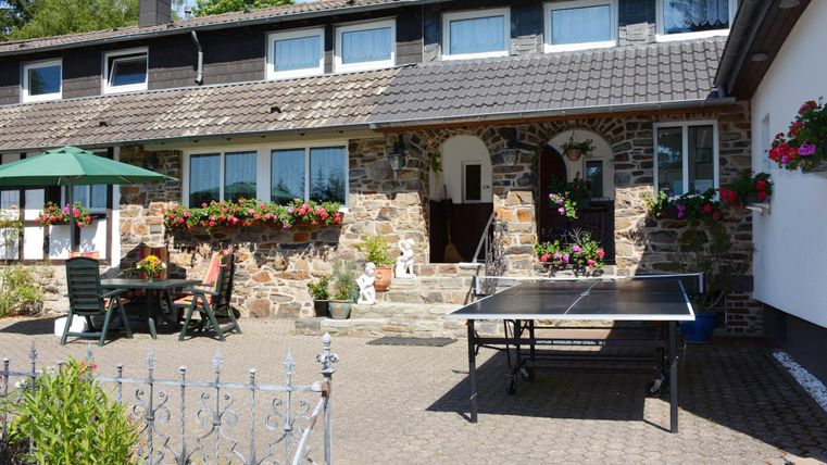 Een gezellig huis met een mooie binnenplaats en bloeiende bloemen. Op de voorgrond staat een tafeltennistafel onder een parasol.