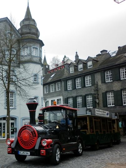 A small tourist train is standing on a paved square. In the background, historical buildings can be seen.