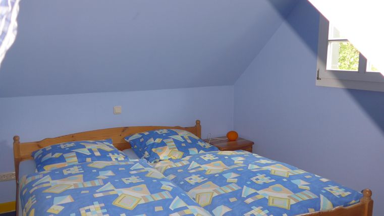 A cozy bedroom with a double bed and blue wall color. The bed has colorful bedding, and there is a window that lets in daylight.
