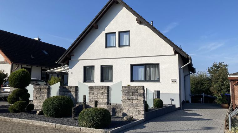 Een modern, twee verdiepingen tellend huis met een witte gevel en grote ramen. De goed onderhouden tuin heeft ronde struiken en een geplaveide oprit.