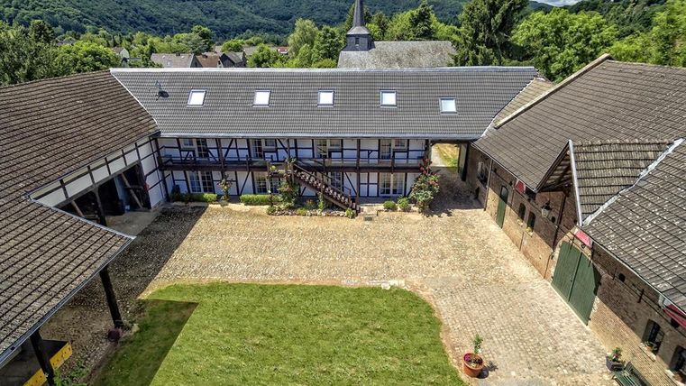 Ein malerischer Innenhof mit modernen Gebäuden und einer grünen Wiese. Im Hintergrund sind bewaldete Hügel und ein Kirchturm sichtbar.