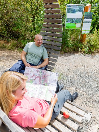 Een paar zit ontspannen op een bank en kijkt naar een kaart. Op de achtergrond zijn informatieborden en groene bomen te zien.