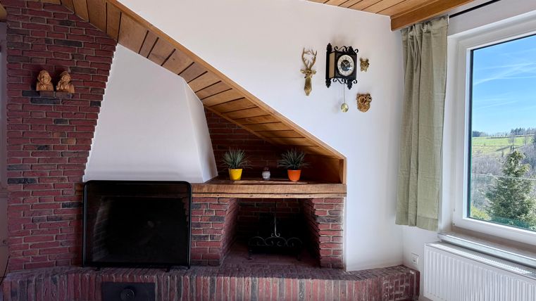 Een gezellige kamer met een bakstenen open haard en scheve houten balken. Een raam met uitzicht op het groen en enkele planten op de vensterbank zorgen voor een uitnodigende sfeer.