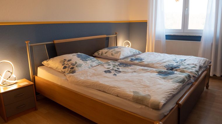 A cozy bedroom with a double bed and bright bedding. The room has a window with curtains and a bedside table with a lamp.