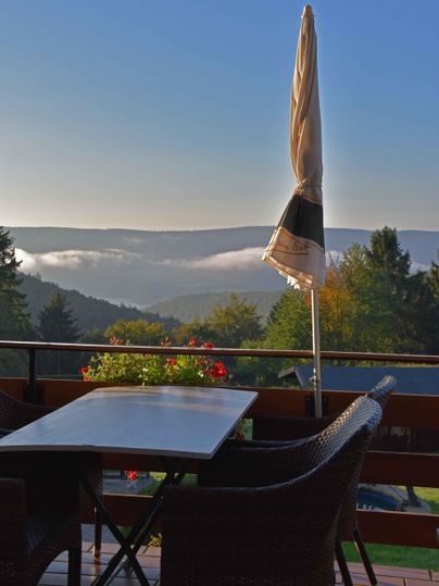 A cozy terrace with a table and chairs, offering a view of the hilly landscape. In the background, fog and trees are visible against a clear sky.