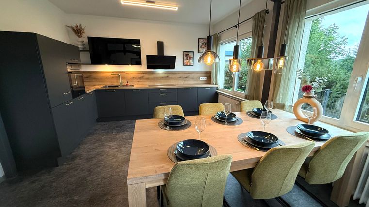 Een moderne keuken met een grote houten tafel en stijlvolle stoelen. Grote ramen zorgen voor veel daglicht en een uitzicht op het groen.
