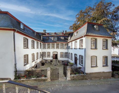 Ein historisches Gebäude mit weißer Fassade und roten Akzenten. Vor dem Gebäude befindet sich ein kleiner Innenhof mit Pflanzen.