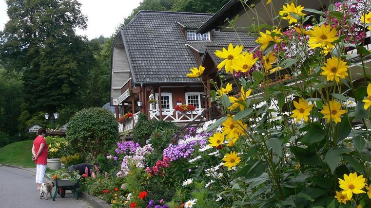 Ein charmantes Haus mit einem Garten voller bunter Blumen. Eine Person geht mit einem Hund und einer Schubkarre vorbei.