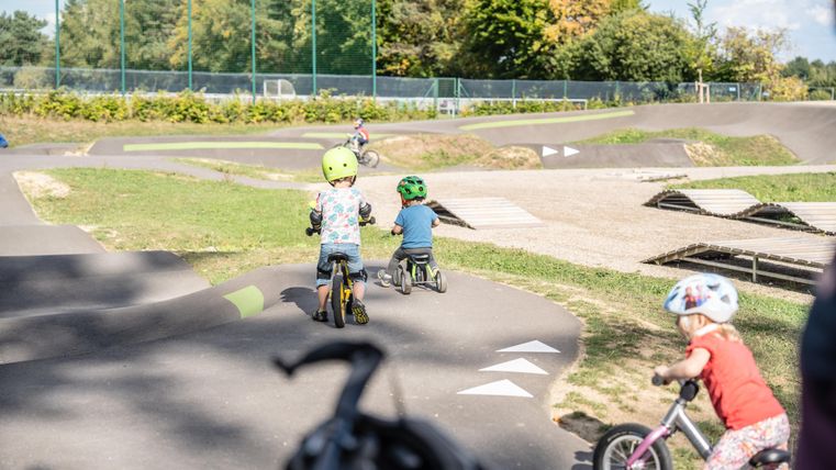 Children are riding their bicycles in a playground. The surroundings are green and sunny, perfect for playing and cycling.