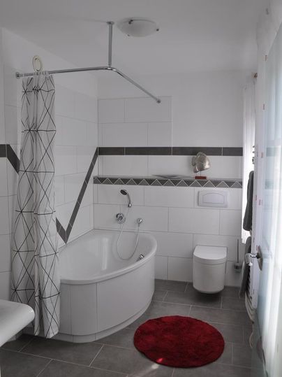 Ein modernes Badezimmer mit einer Badewanne und einer Dusche. Der Boden ist mit grauen Fliesen ausgelegt, und ein rotes Badezimmerteppichstück fügt einen Farbtupfer hinzu.