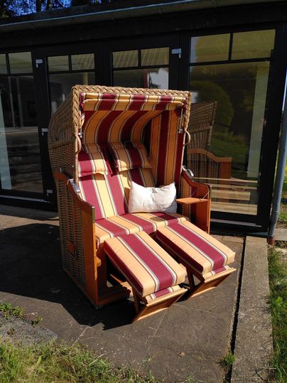 A cozy beach chair with striped cushions is sitting on a paved area. In the background, large windows and a green garden can be seen.