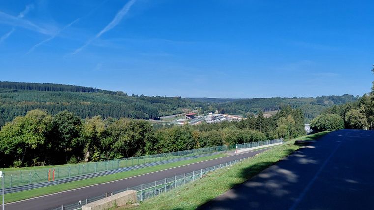 A picturesque view of a green landscape with a clear blue sky. In the foreground, there is a road that winds through the nature.